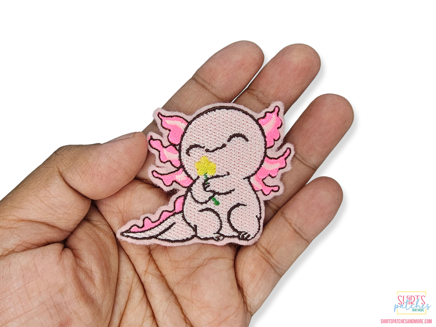 Hand holding a cute pink axolotl embroidered patch to show size. The axolotl is smiling and holding a yellow flower.