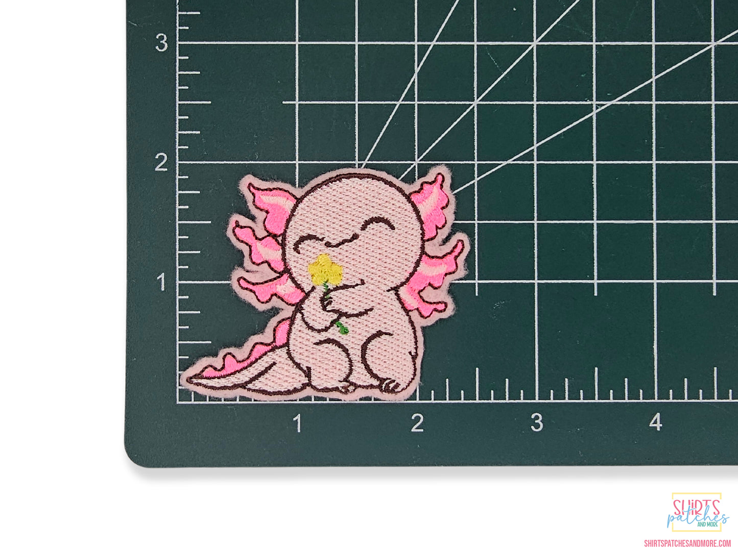 Pink kawaii axolotl embroidered patch shown on a green cutting mat with grid lines to demonstrate its 2.5-inch width and size.