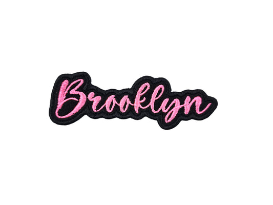 Brooklyn custom name patch by Shirts Patches And More, embroidered in pink script with a black border on a white background.