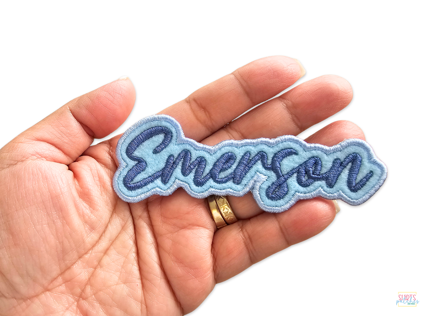 Hand holding a personalized Emerson name patch featuring dark blue script lettering on a light blue embroidered background.