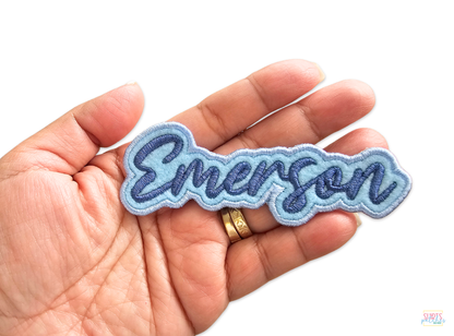 Hand holding a personalized Emerson name patch featuring dark blue script lettering on a light blue embroidered background.