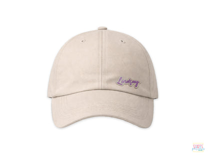 A beige baseball cap featuring a custom 'Lindsay' embroidered name patch with purple script text, shown on a white background.