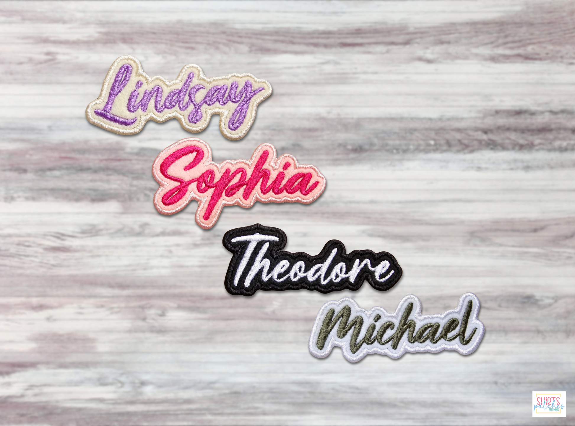 Four personalized embroidered name patches in different colors (purple, pink, black, and white) with script lettering displayed on a light wood background.