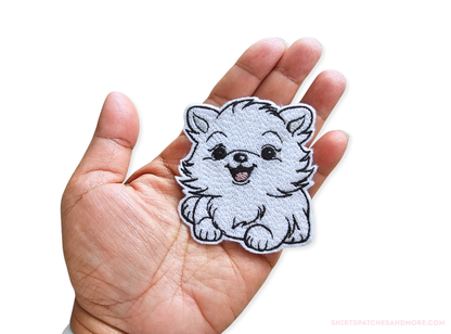 Embroidered white Pomeranian puppy patch held in a hand to show scale, approximately 2.7 inches wide by 3 inches tall, featuring a happy expression and fluffy fur detail on a white background.