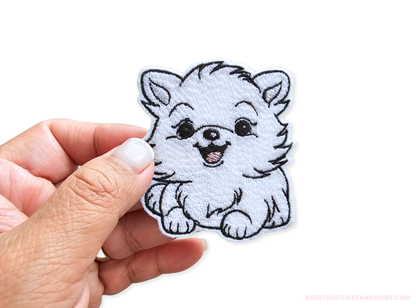 Embroidered white Pomeranian puppy patch held in a hand to show scale, approximately 2.7 inches wide by 3 inches tall, featuring a happy expression and fluffy fur detail on a white background.