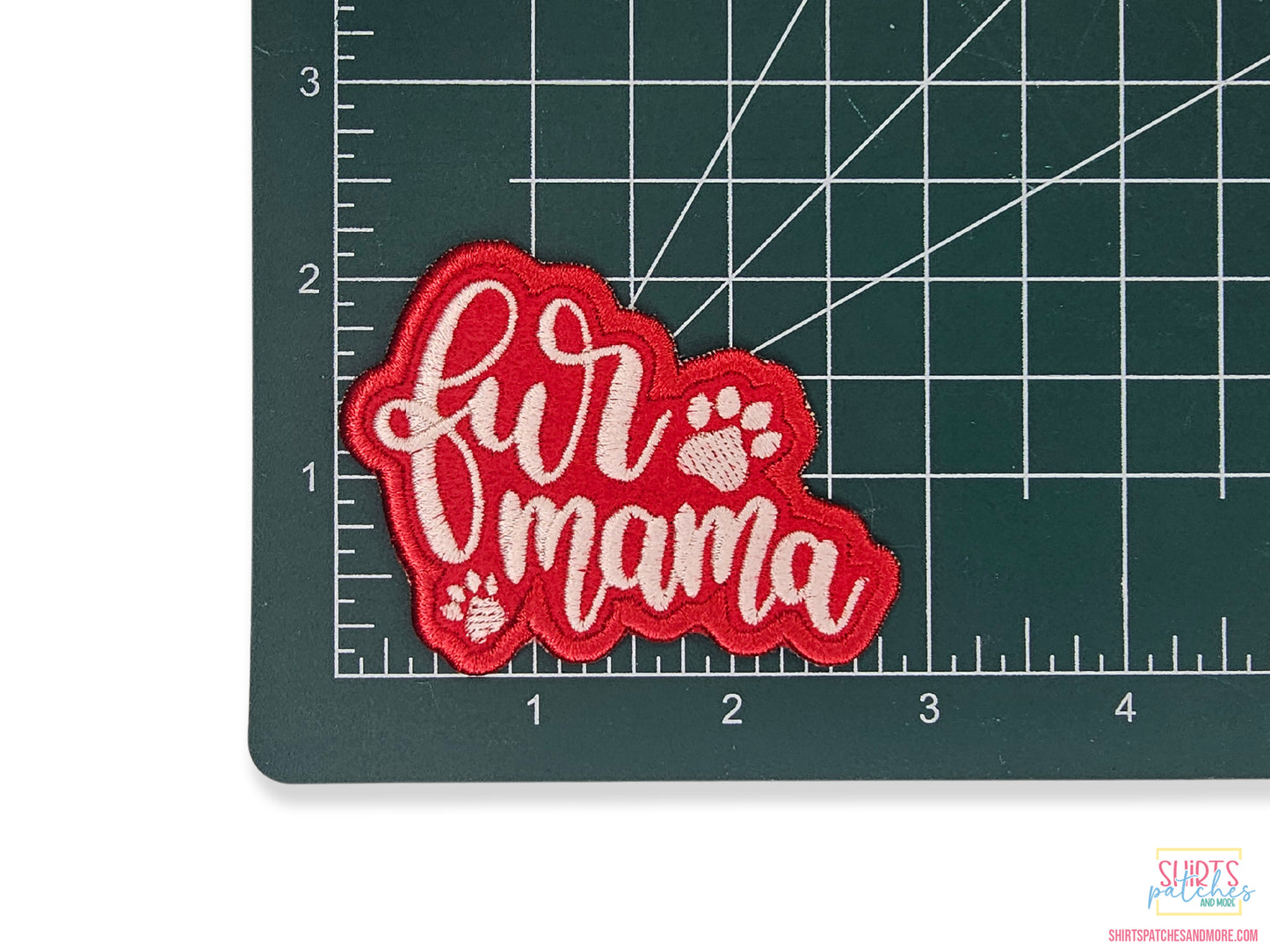 A red fabric patch featuring the words "fur mama" in white cursive with two paw prints, placed on a green gridded background showing the patch is approximately 2.8 inches wide.