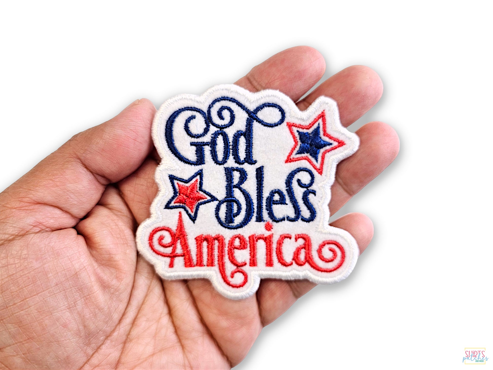 A person's hand holding a high-quality "God Bless America" embroidered patch with red and navy blue script and patriotic stars against a white background.