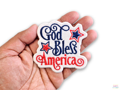 A person's hand holding a high-quality "God Bless America" embroidered patch with red and navy blue script and patriotic stars against a white background.
