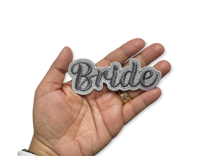 Close-up of a hand holding a "Bride" embroidered patch with silver script lettering and a white border against a white background.