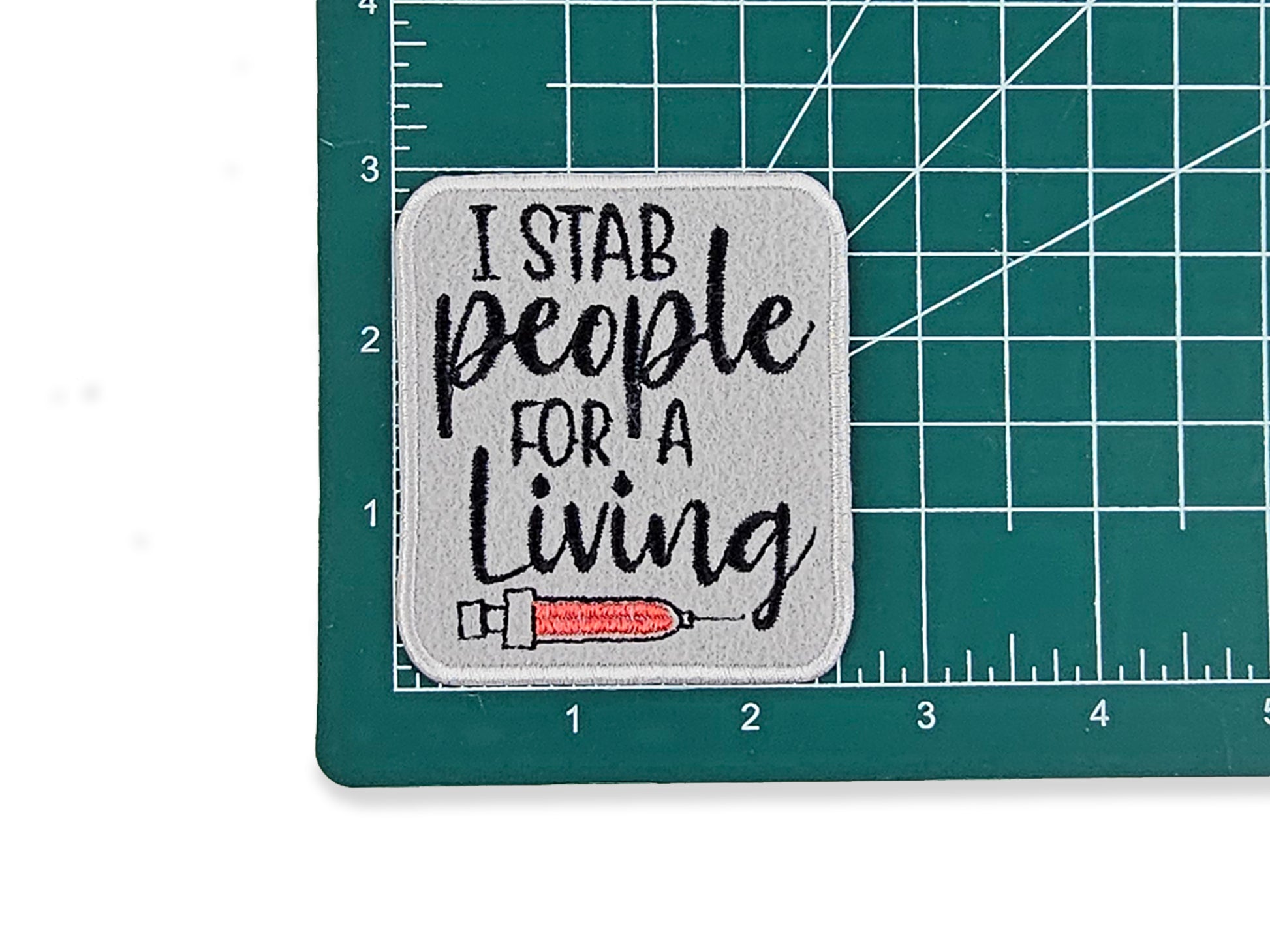 I Stab People for a Living" funny nurse patch on a green grid cutting mat to show the 2.6" x 3" size, featuring a gray background and red syringe embroidery.
