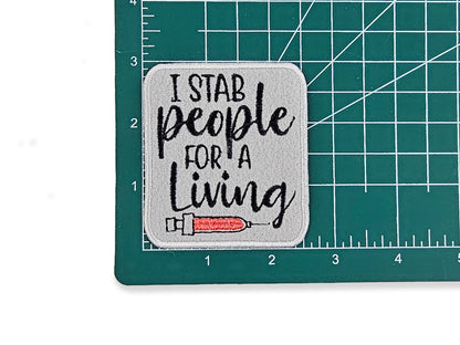 I Stab People for a Living" funny nurse patch on a green grid cutting mat to show the 2.6" x 3" size, featuring a gray background and red syringe embroidery.