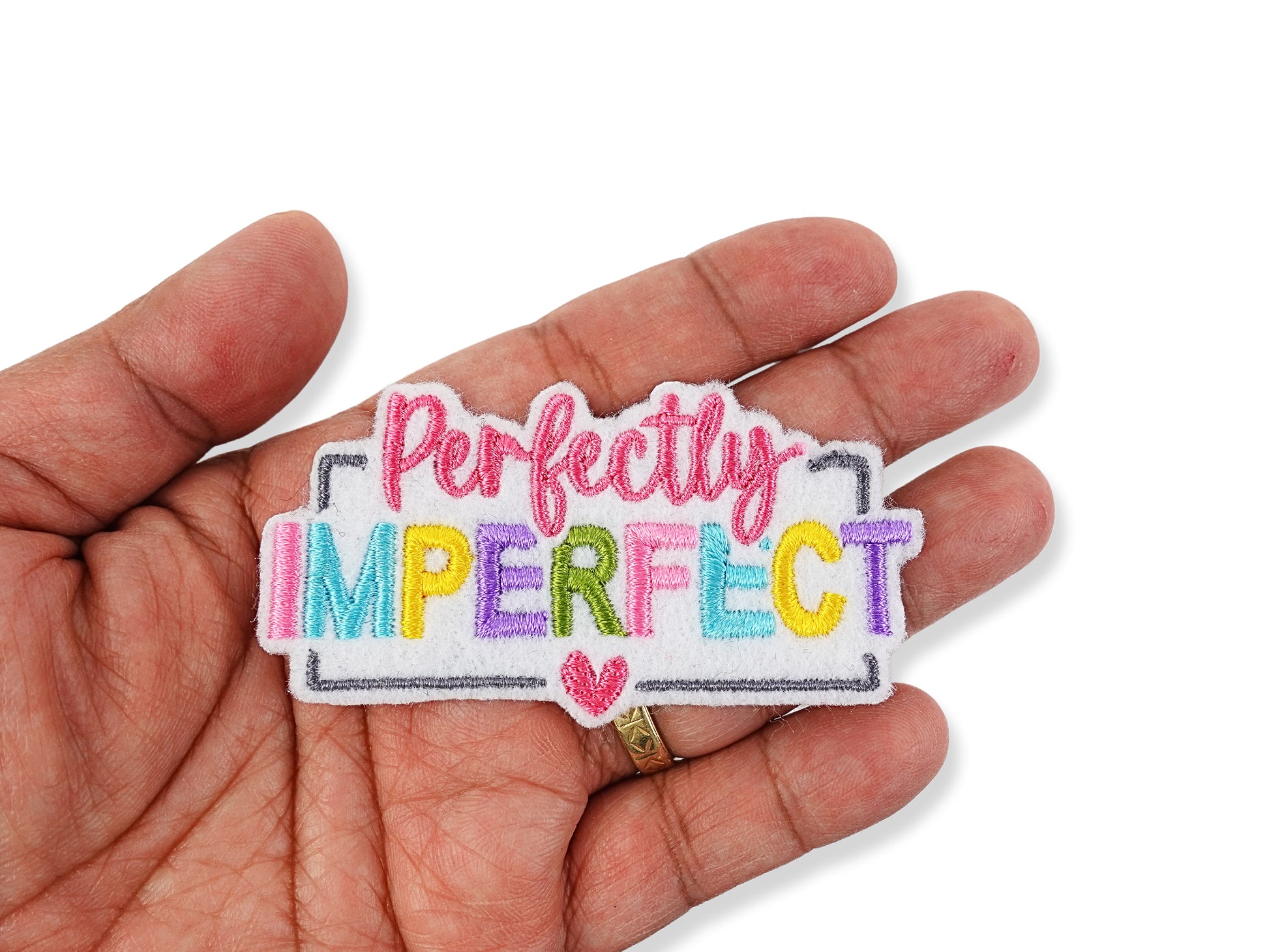 Hand holding a "Perfectly Imperfect" embroidered patch to show scale, featuring multi-colored rainbow lettering, a pink script font, and a small heart detail on a white background.
