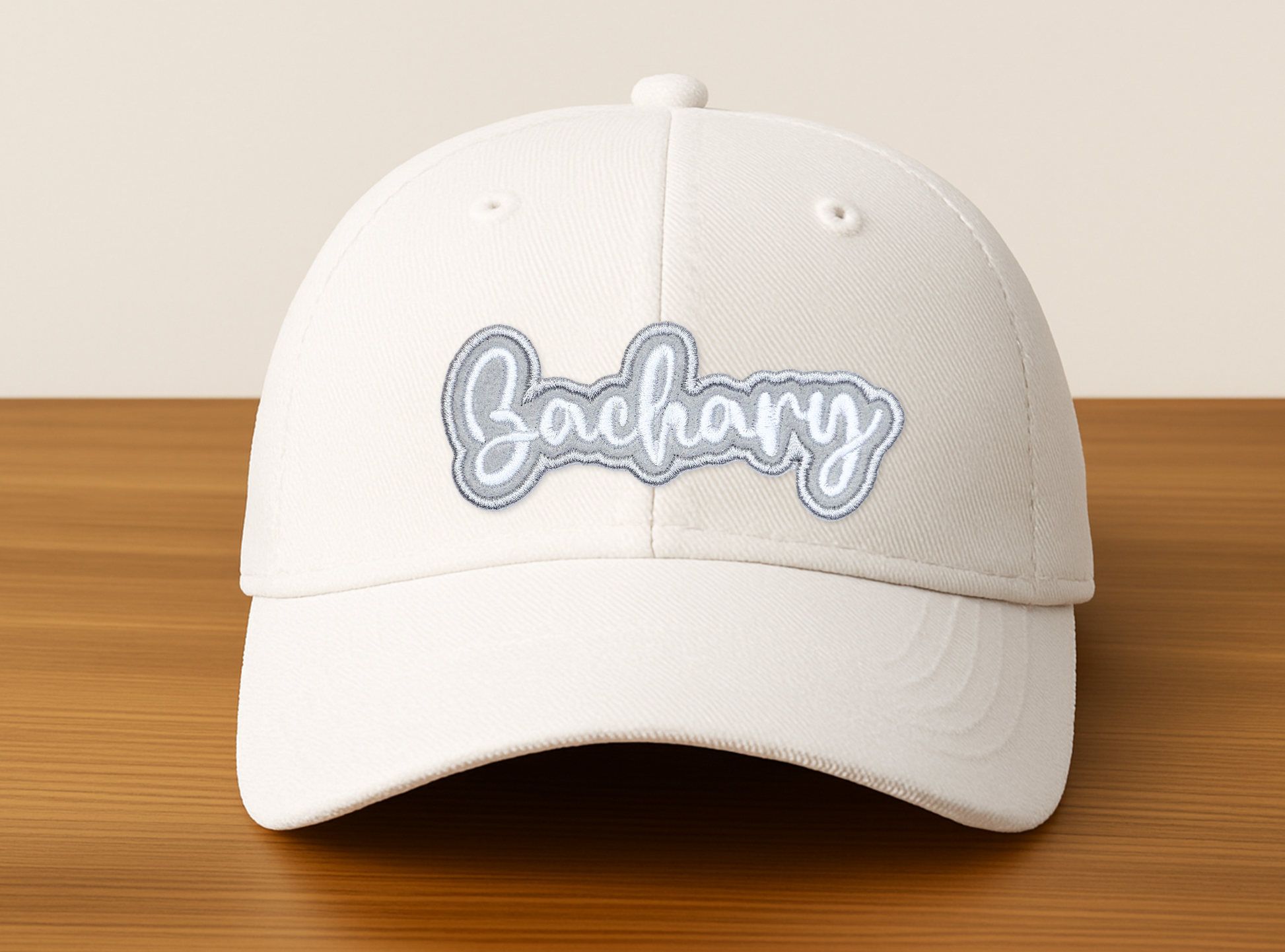 White embroidered name patch that says “Zachary” applied to a cream baseball hat, showing a personalized custom iron-on patch style on headwear.