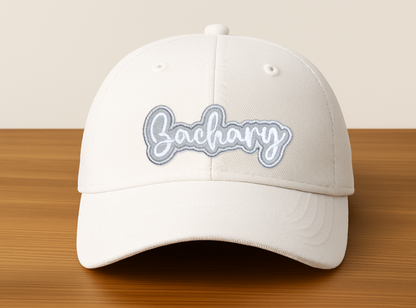 White embroidered name patch that says “Zachary” applied to a cream baseball hat, showing a personalized custom iron-on patch style on headwear.