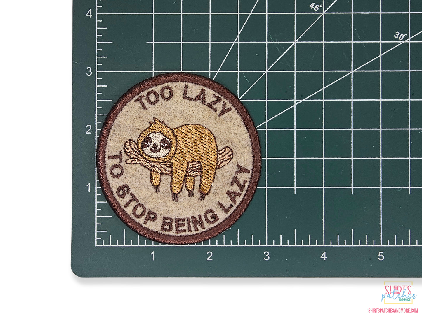 3-inch "Too Lazy to Stop Being Lazy" sloth embroidered patch shown on a green metric cutting mat to demonstrate size and high-quality tan and brown stitching.