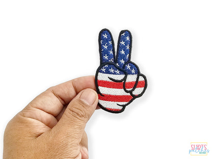 Hand holding a patriotic peace sign patch featuring a red, white, and blue American flag stars and stripes design against a white background.