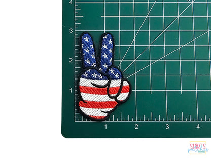 Embroidered peace sign hand patch with American flag stars and stripes design, shown on a green self-healing cutting mat with inch measurements for scale.
