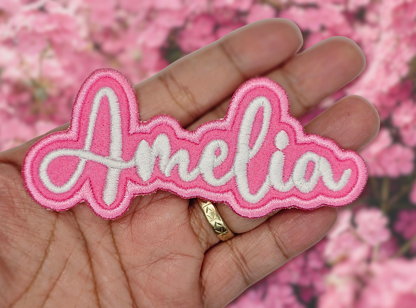 Pink and white 'Amelia' name patch held in a hand with a pink floral background