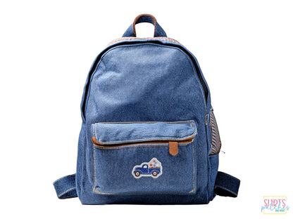 A blue denim backpack with a small front pocket featuring a blue patriotic vintage truck embroidered patch on a white background.