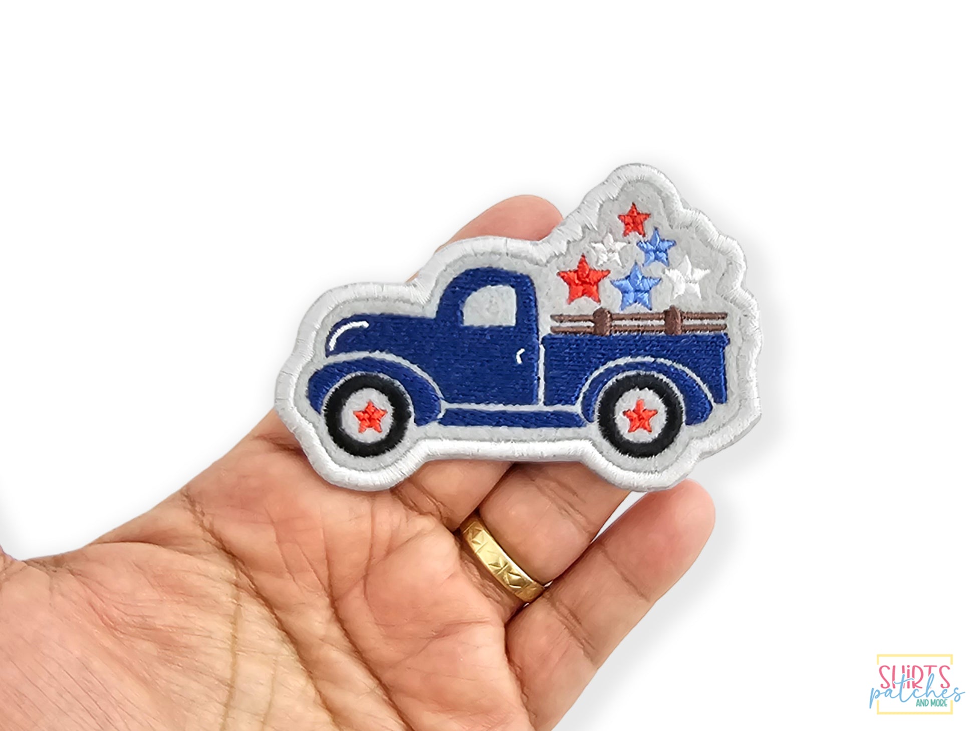 Close-up of a blue vintage pickup truck embroidered patch with stars on the wheels and a star-filled truck bed, held in a person's hand to show its relative size.