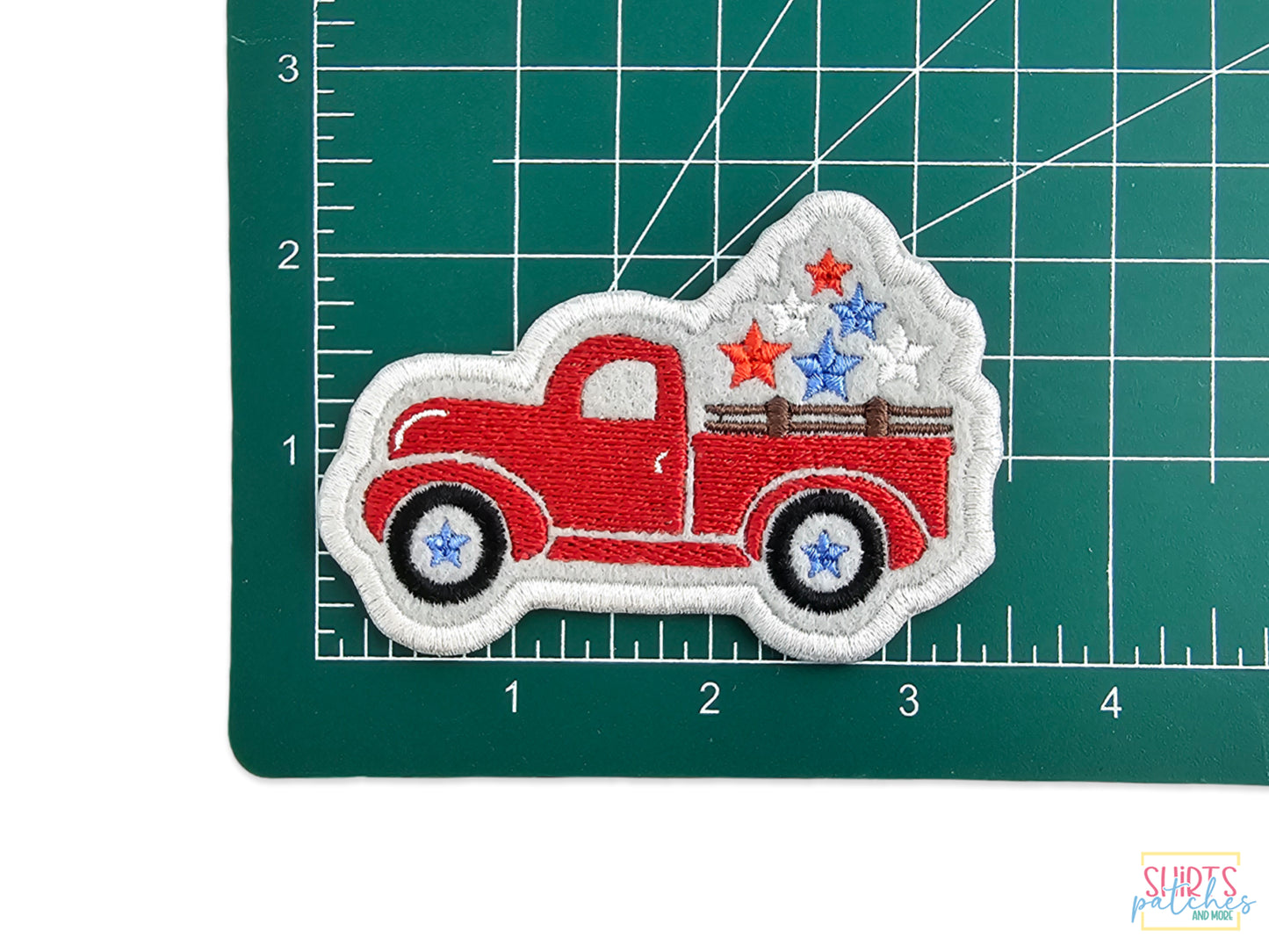 Red embroidered vintage pickup truck patch with white, blue, and red stars in the truck bed, shown on a green measuring mat.