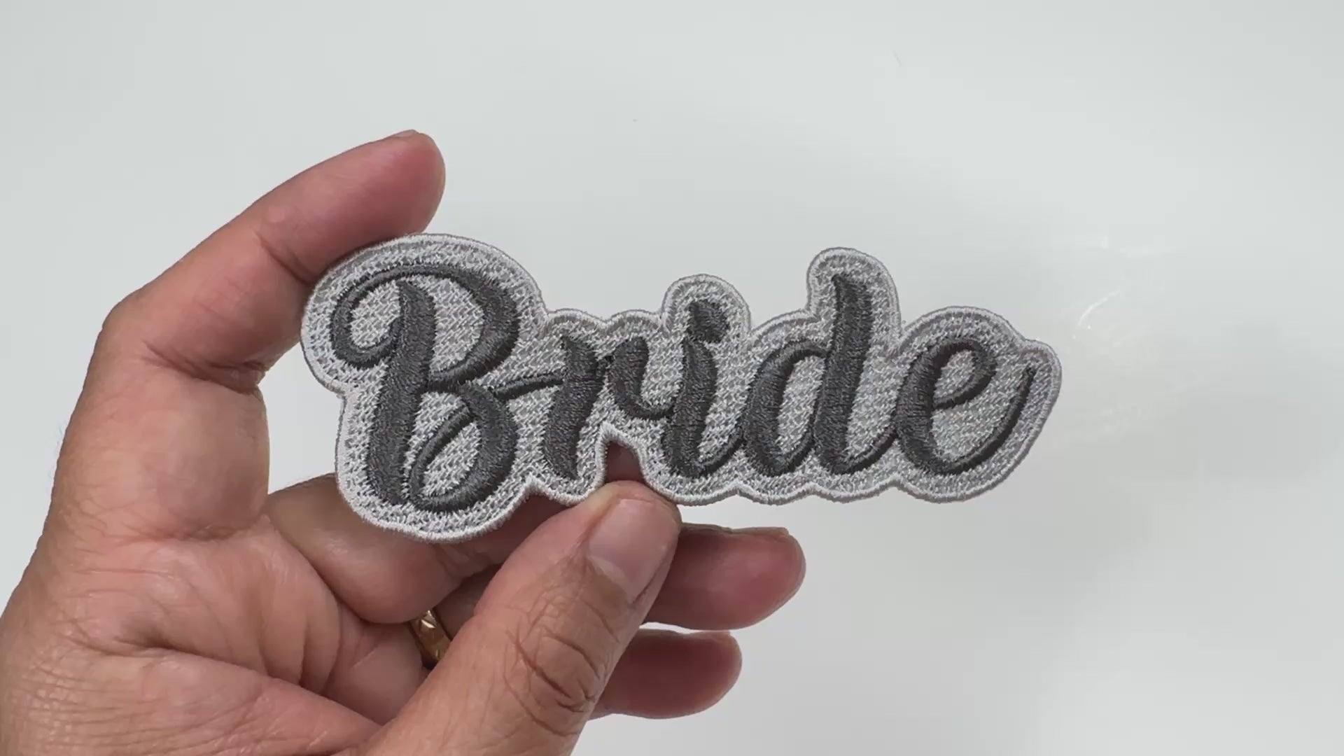 Handheld close-up video of a "Bride" embroidered patch with silver-gray script lettering and a dark border, showcasing the high-quality stitching and texture.