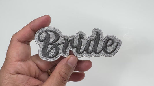 Handheld close-up video of a "Bride" embroidered patch with silver-gray script lettering and a dark border, showcasing the high-quality stitching and texture.