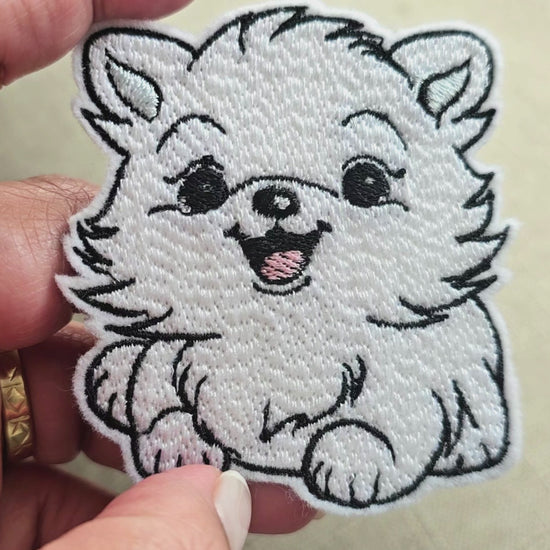 Video showing a hand holding a white embroidered Pomeranian puppy patch, highlighting the detailed stitching and fluffy texture against a neutral background