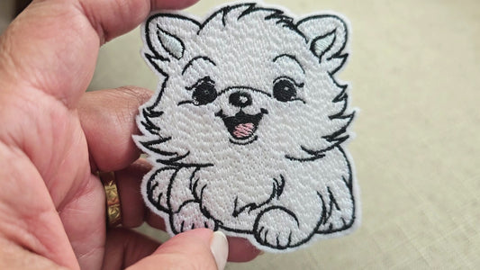 Video showing a hand holding a white embroidered Pomeranian puppy patch, highlighting the detailed stitching and fluffy texture against a neutral background