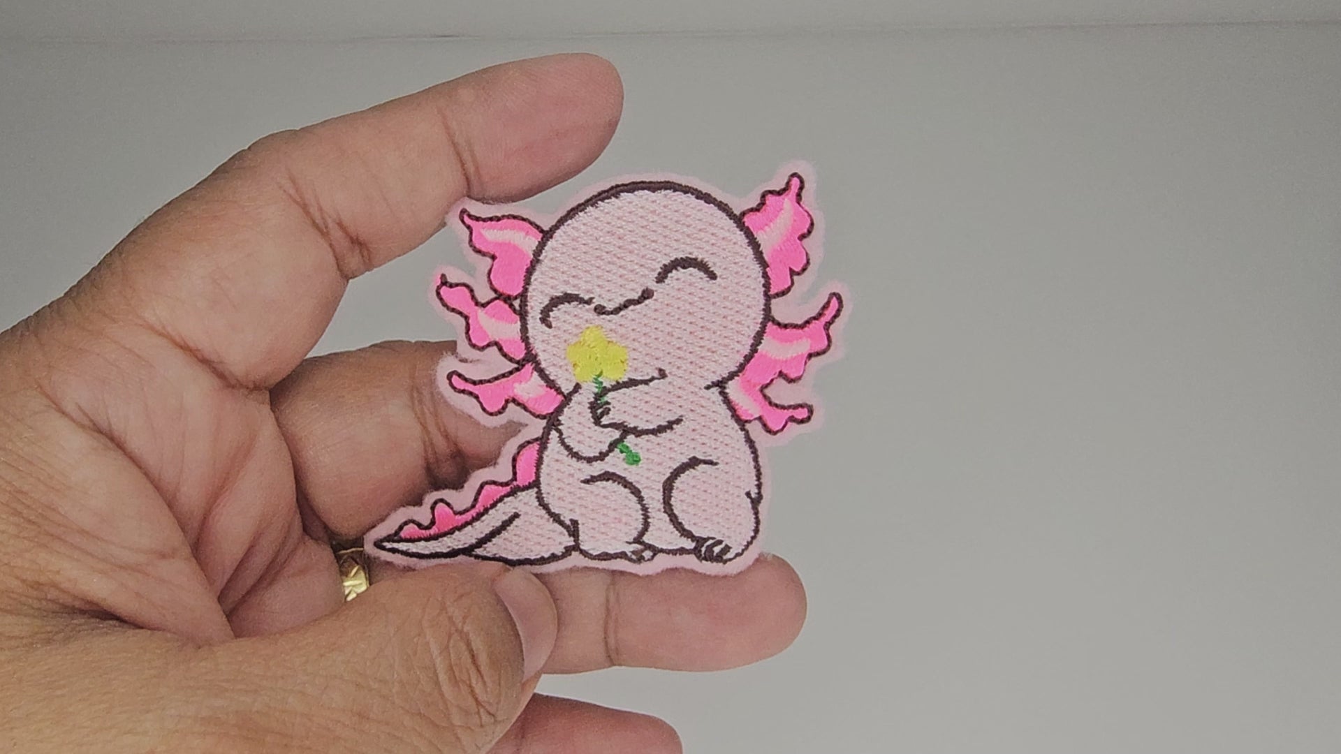 Video clip of a hand holding the cute pink axolotl embroidered patch to demonstrate size and stitching quality.