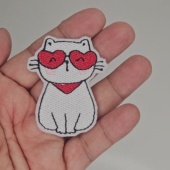 Video showing details of white cat embroidered patch with red heart sunglasses and scarf held in hand