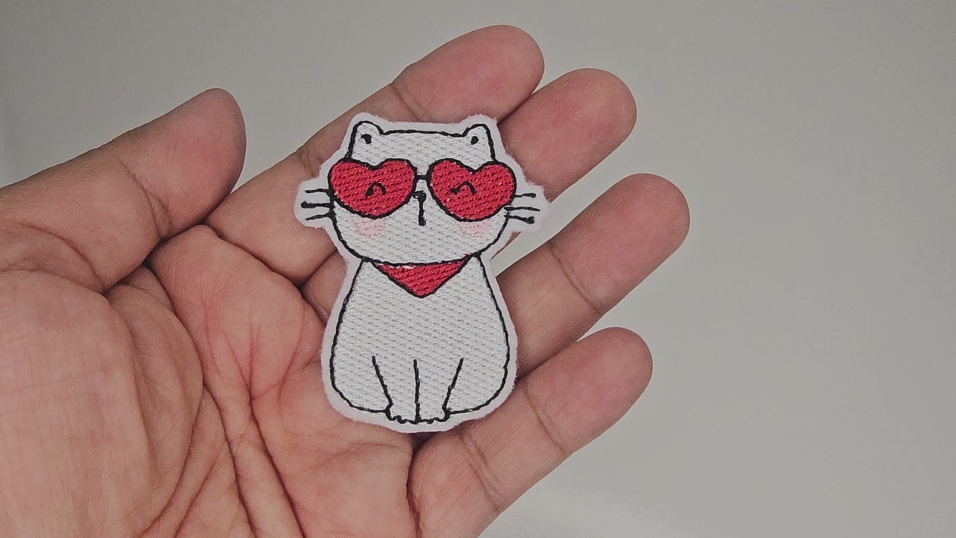 Video showing details of white cat embroidered patch with red heart sunglasses and scarf held in hand