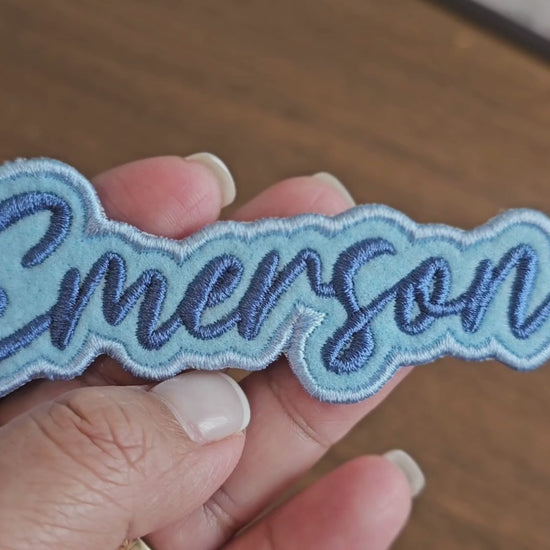 Video showing a close-up of a custom Emerson name patch with dark blue script lettering on a light blue embroidered background being held by a hand.