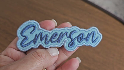 Video showing a close-up of a custom Emerson name patch with dark blue script lettering on a light blue embroidered background being held by a hand.