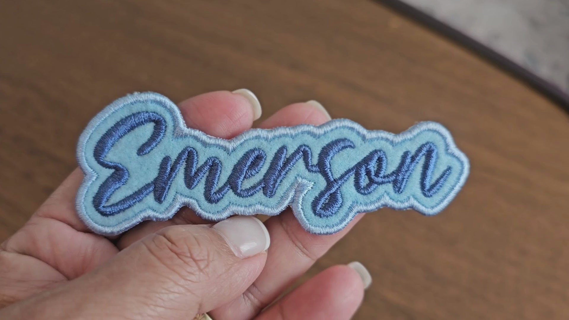 Video showing a close-up of a custom Emerson name patch with dark blue script lettering on a light blue embroidered background being held by a hand.