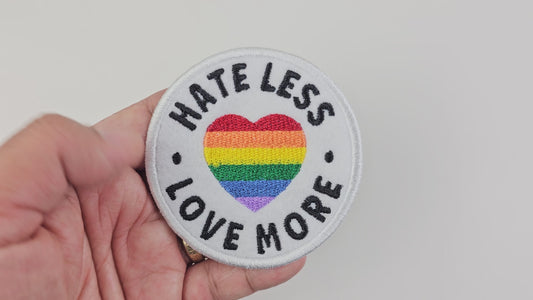 Video showing a hand holding a white circular embroidered patch with a rainbow pride heart and the text "Hate Less Love More" in bold black letters.