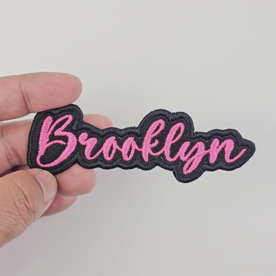 Video showcasing the Brooklyn custom embroidered patch, held in hand, highlighting the pink script and black border.