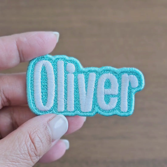 Video showing a close-up of the 'Oliver' custom embroidered name patch with light blue border and white script lettering, held in a hand for scale and detail.