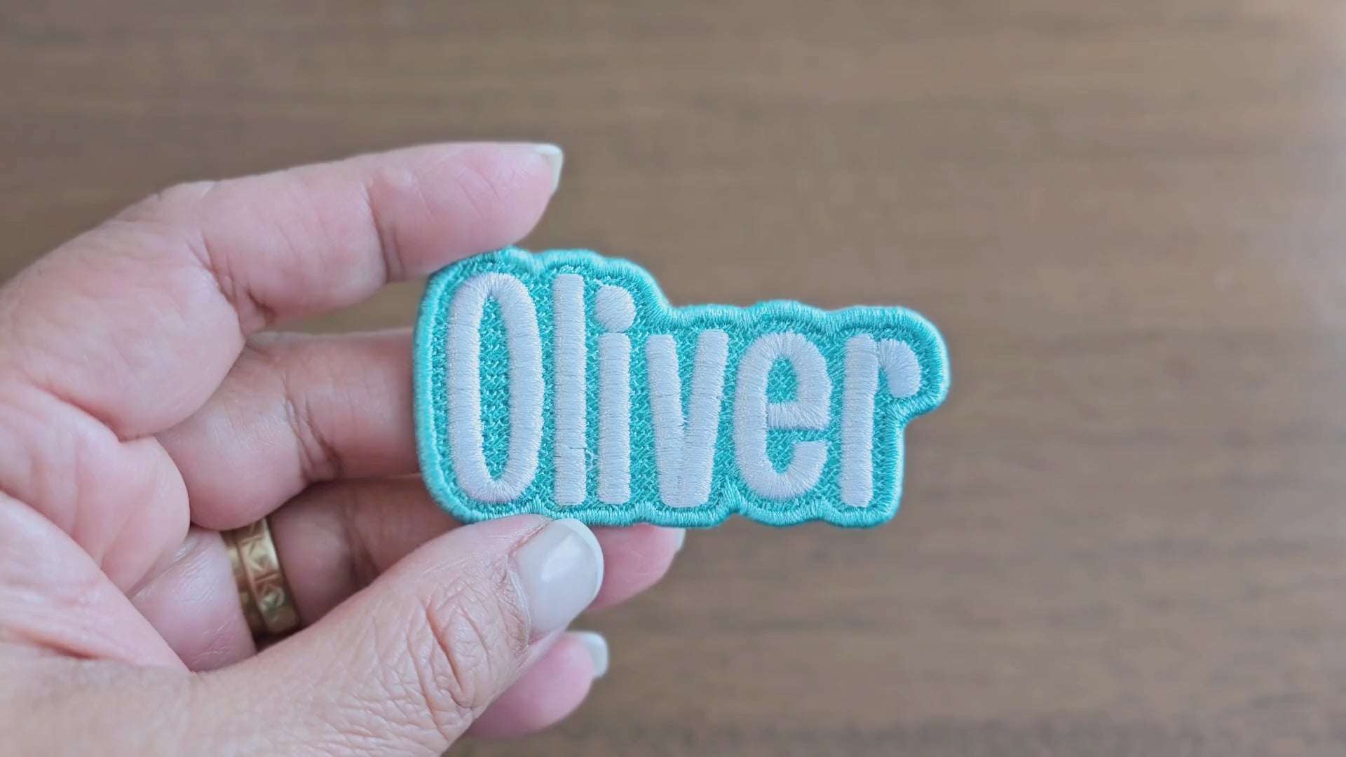 Video showing a close-up of the 'Oliver' custom embroidered name patch with light blue border and white script lettering, held in a hand for scale and detail.