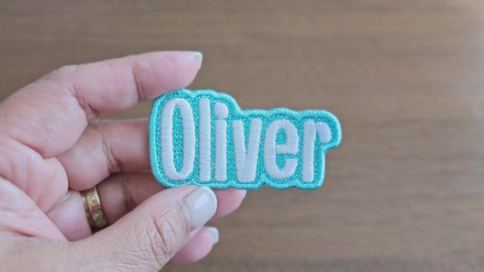 Video showing a close-up of the 'Oliver' custom embroidered name patch with light blue border and white script lettering, held in a hand for scale and detail.