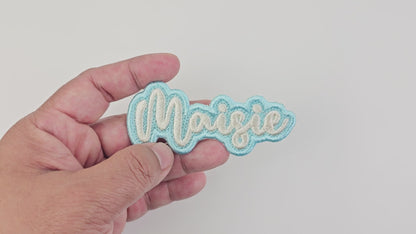 Video showing a close-up of the “Maisie” custom embroidered name patch being held and rotated in hand, highlighting the satin stitching, outline, and color details—personalized iron-on, sew-on, stick-on, and Velcro patch option