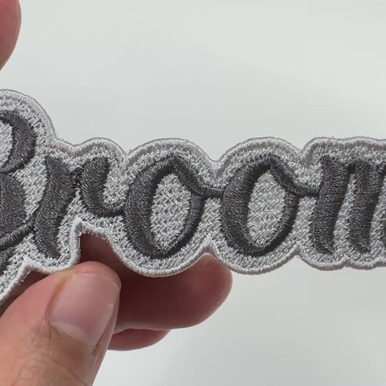Handheld video showing a high-quality 'Groom' embroidered patch with dark grey script lettering and a light grey bordered edge, demonstrating the detailed stitching.
