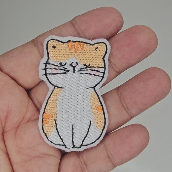 Video of hand holding a white and orange tabby cat embroidered patch to show size and quality