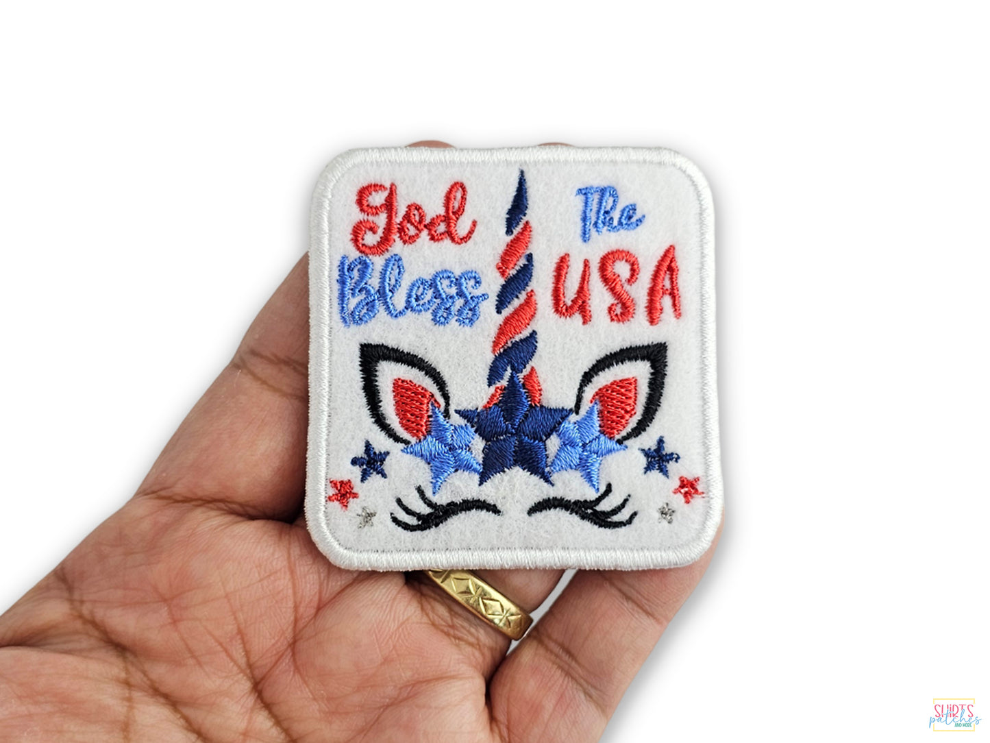 A person's hand holding a white embroidered patch featuring a unicorn face and the words 'God Bless The USA' in red and blue script.